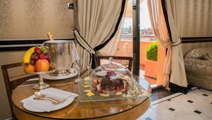 A dining table with a fruit bowl, a glass of water, a dessert under a glass dome, and a view of the outdoors through a window with curtains.