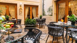 Un área de comedor con mesas redondas y sillas de mimbre negro, rodeada de plantas en macetas y un mural decorativo.