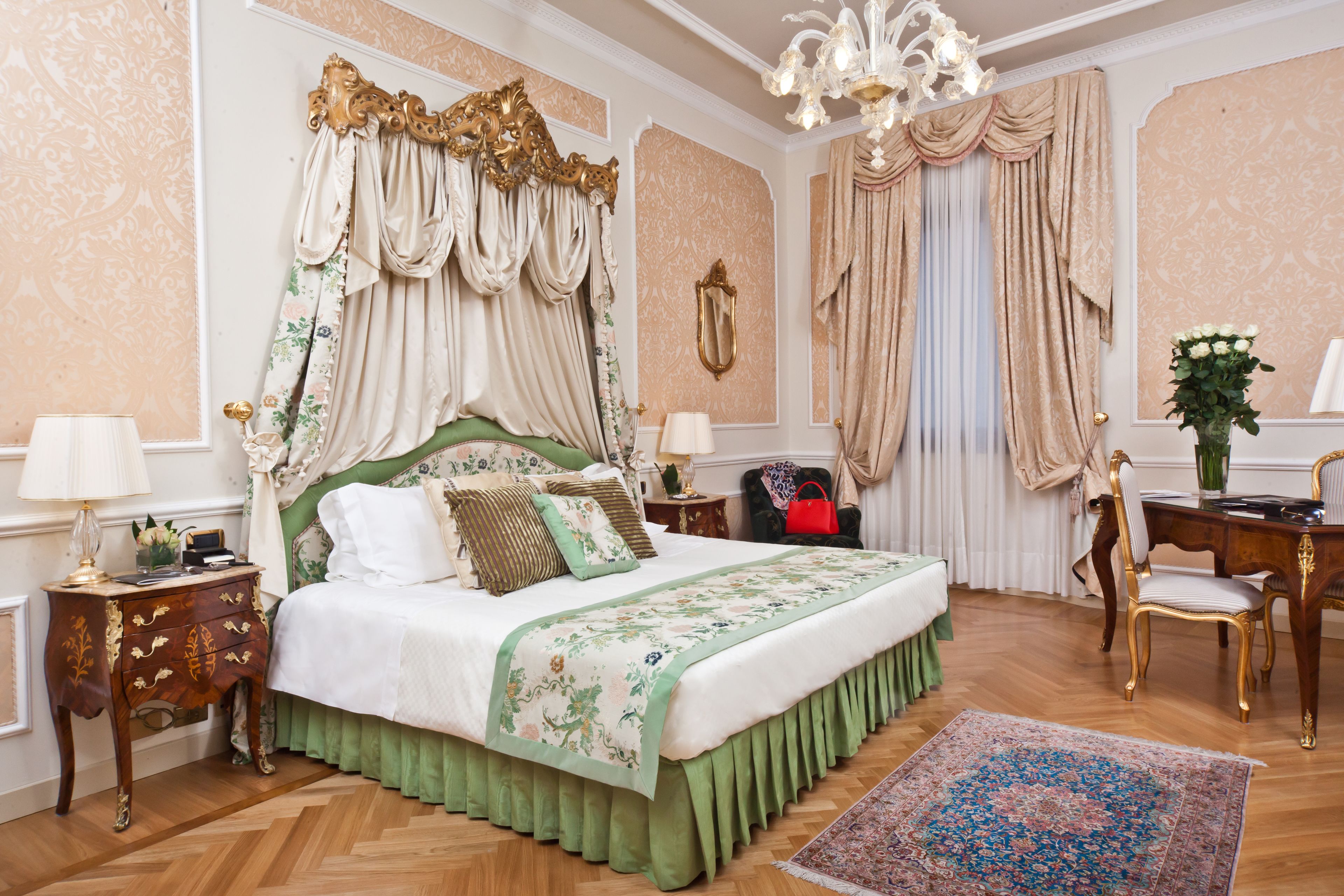 A classic bedroom with a large bed, ornate curtains, a chandelier, and a green upholstered chair.