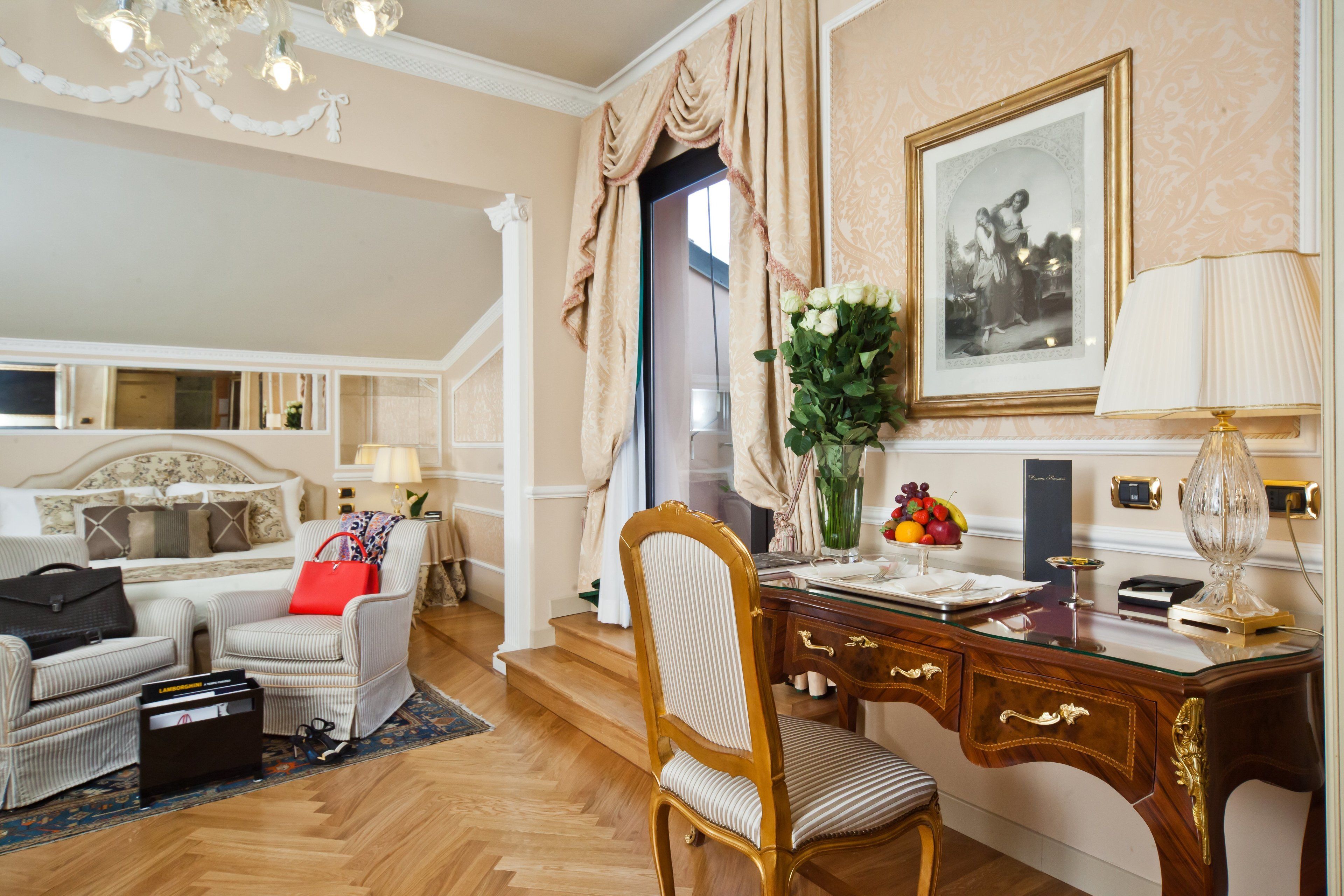 Una habitación de hotel elegante con cama, una zona de estar con una silla y una mesa, un escritorio con lámpara y un cuadro en la pared.