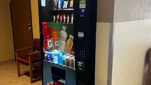 Máquina expendedora con diversas bebidas y aperitivos, ubicada en una sala con suelo de baldosas y una silla de madera.