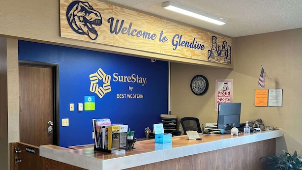 Un mostrador de recepción con un letrero que dice 'Welcome to Glendale', el logotipo de 'SureStay by Best Western', un reloj y una bandera estadounidense.