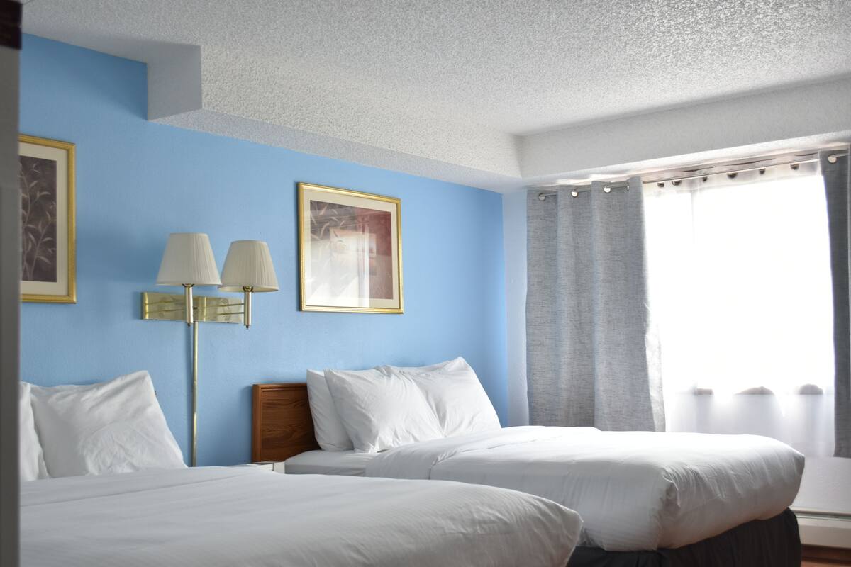A hotel room with two beds, a nightstand, a lamp, and a window with curtains.