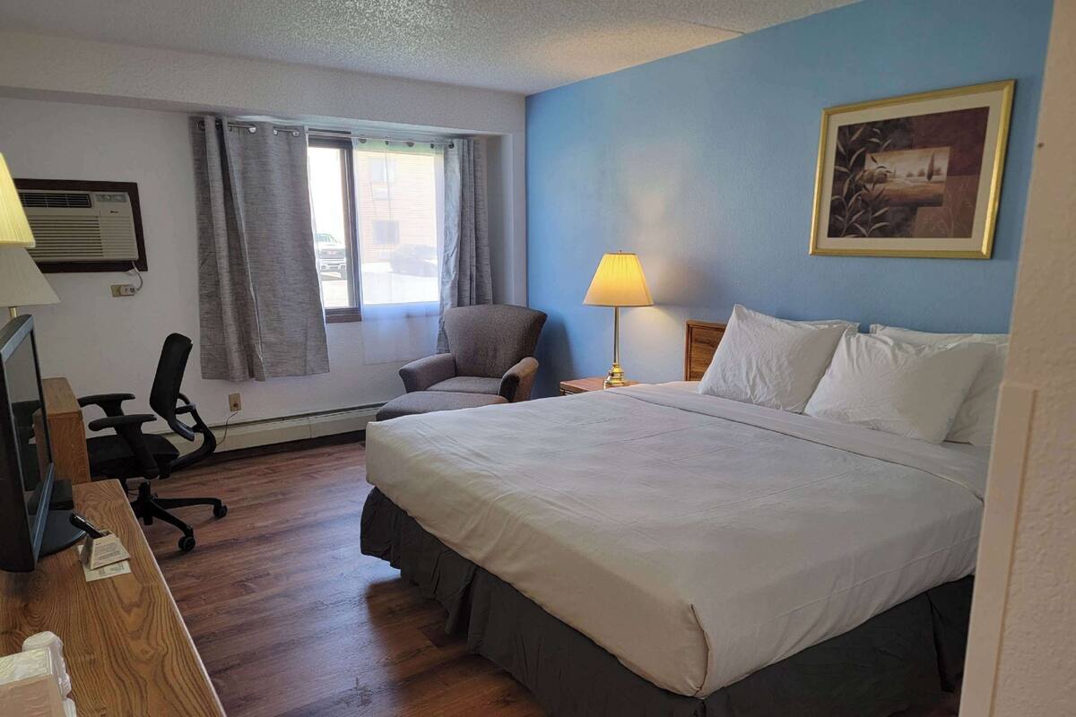 A hotel room with a large bed, a desk with a computer, a chair, a window with curtains, and a painting on the wall.