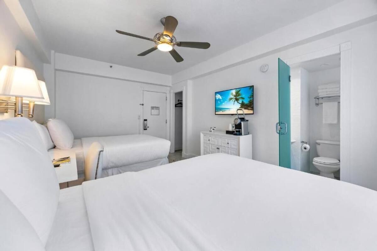 A hotel room with two beds, a ceiling fan, a TV, a minibar, and a bathroom with a toilet and towel rack.