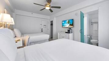 Quarto de hotel com duas camas, ventilador de teto, televisão, frigobar e banheiro com vaso sanitário e porta-toalhas.