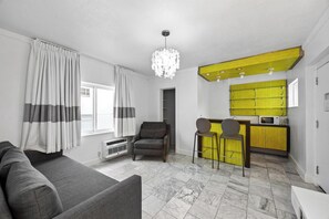 A modern living room with a grey sofa, a dark armchair, and a yellow kitchen area with a microwave.