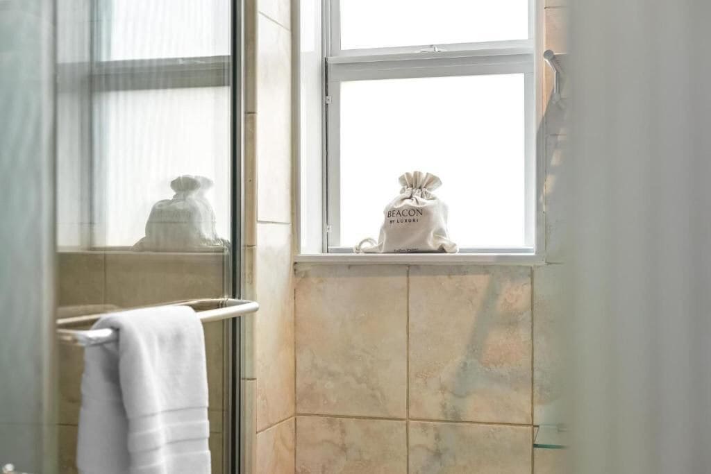 A bathroom with a window, a towel rack, and a bag labeled "BEACON BY LUXURY" on the windowsill.