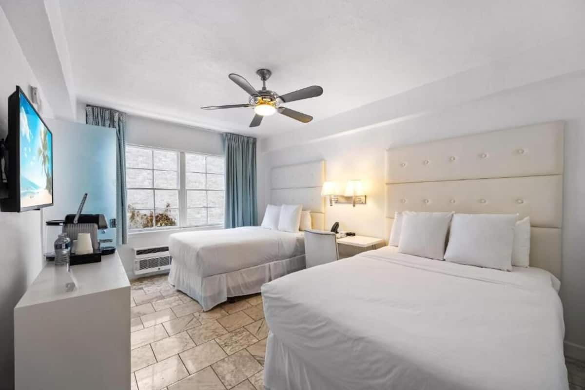 A hotel room with two beds, a ceiling fan, a TV, a desk with a lamp, and a window with curtains.