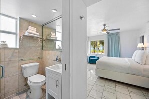 A modern bathroom with a toilet, sink, and mirror. There is a bed with a ceiling fan in the adjacent room.