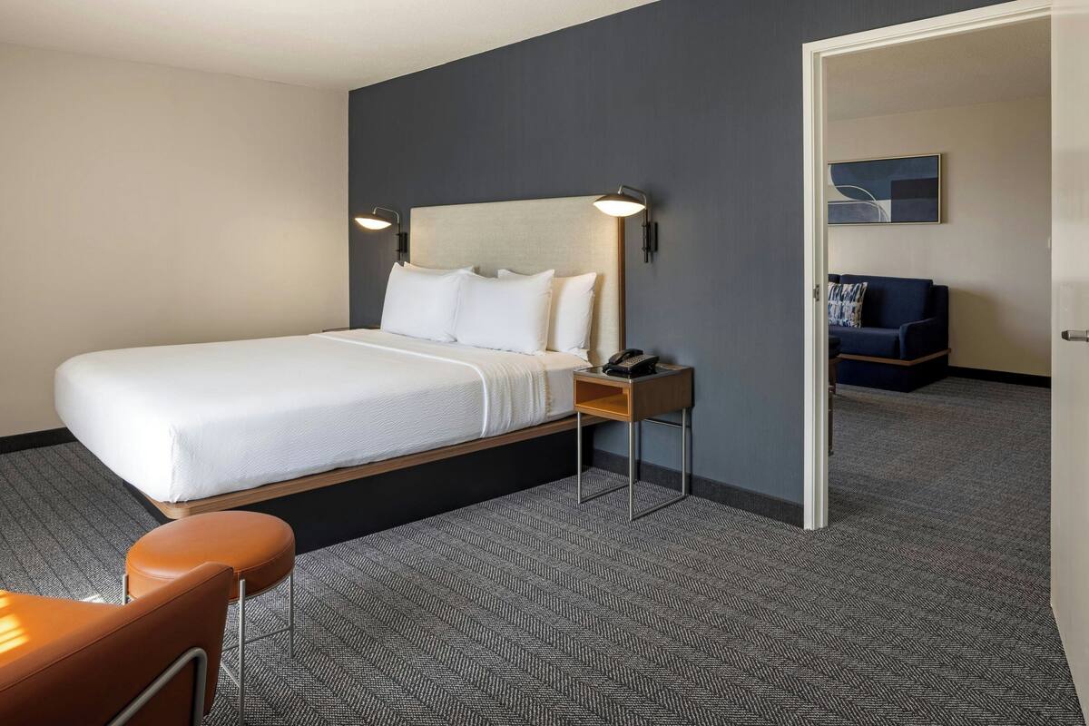 A hotel room with a bed, a nightstand, a chair, and a door leading to another room.