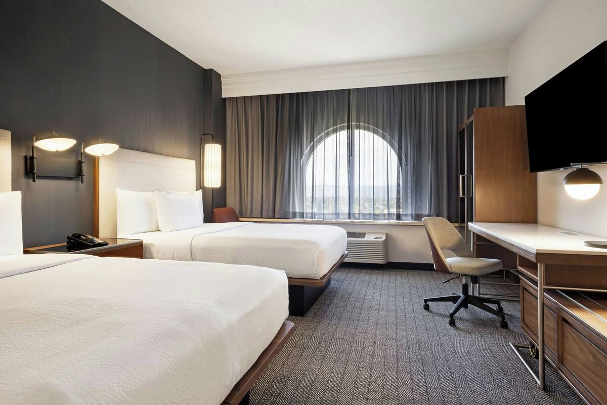 A hotel room with two beds, a desk, a chair, a TV, and a large window with curtains.