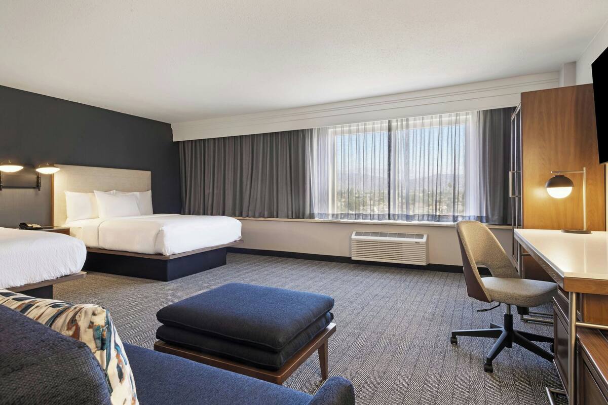 A hotel room with a bed, a desk, a chair, and a view of the outdoors through large windows.