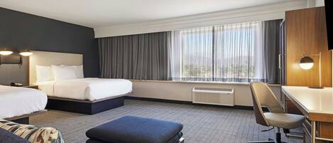 A hotel room with a bed, a desk, a chair, and a view of the outdoors through large windows.