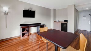 Quarto de hotel moderno com chão de madeira, uma mesa de madeira escura, duas cadeiras, uma TV de tela plana na parede, uma pequena cozinha compacta e uma porta com uma placa.