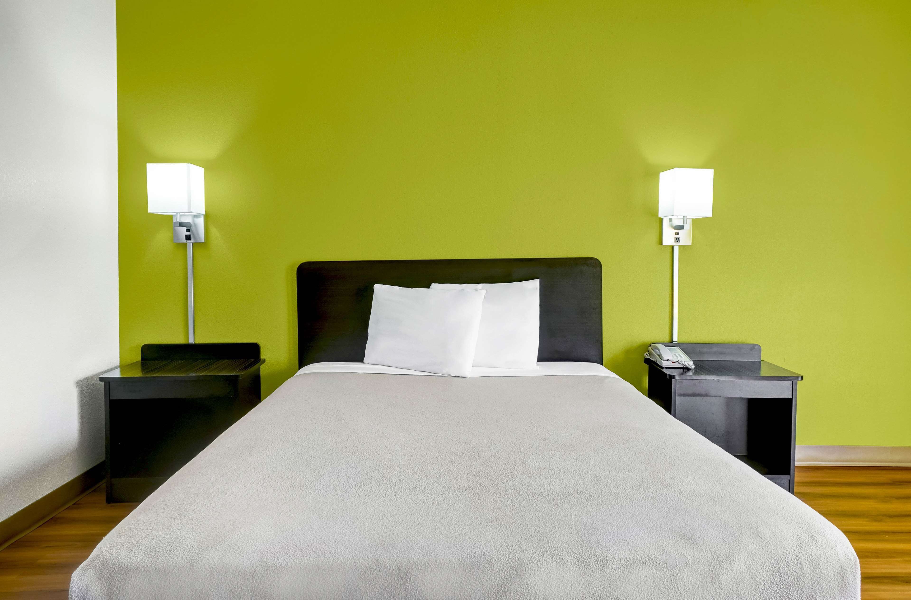 A bed with a white headboard and pillows, flanked by two nightstands with lamps, in a room with a green wall and wooden flooring.
