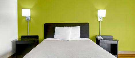 A bed with a white headboard and pillows, flanked by two nightstands with lamps, in a room with a green wall and wooden flooring.