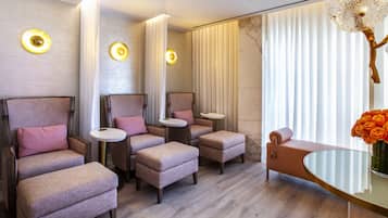 A modern spa room with multiple seating areas, including cushioned chairs and ottomans, a small round table, and a glass table with a vase of flowers.