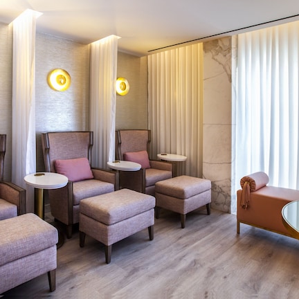 A modern spa room with multiple seating areas, including cushioned chairs and ottomans, a small round table, and a glass table with a vase of flowers.