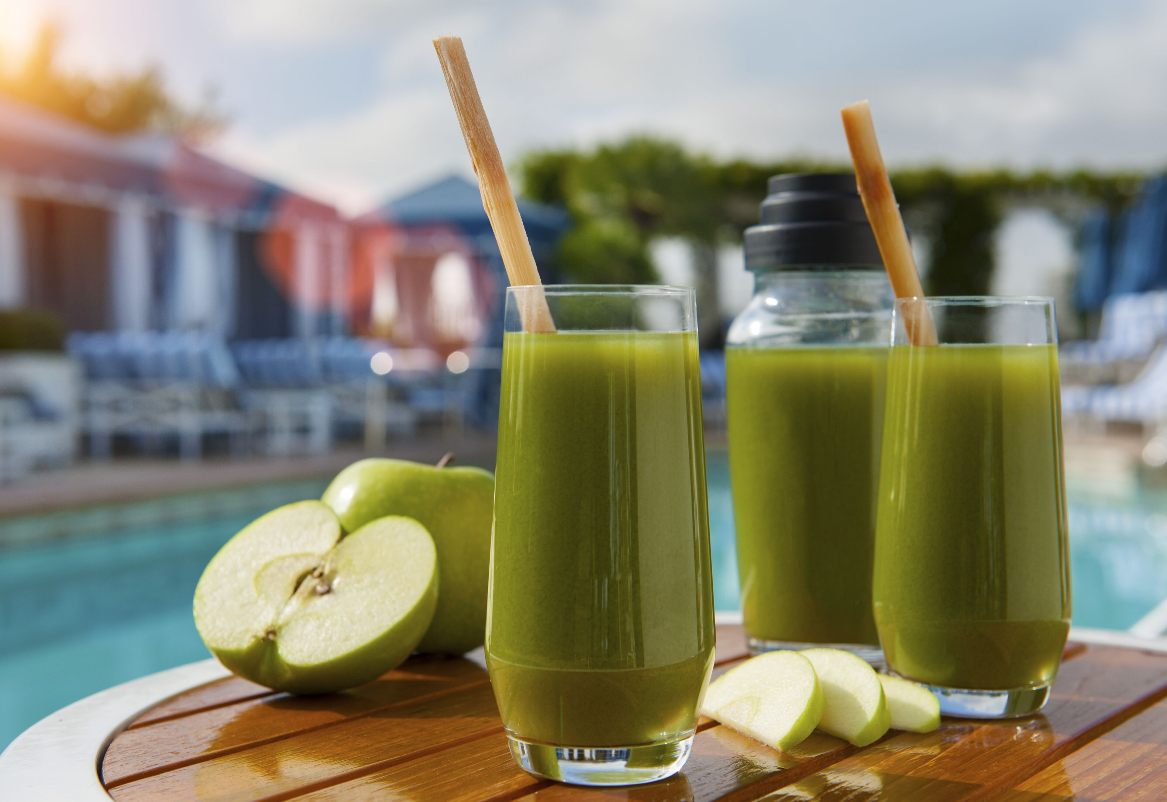 A poolside setting with two glasses of green juice, a half apple, and a bottle of juice, with a pool and lounge chairs in the background.