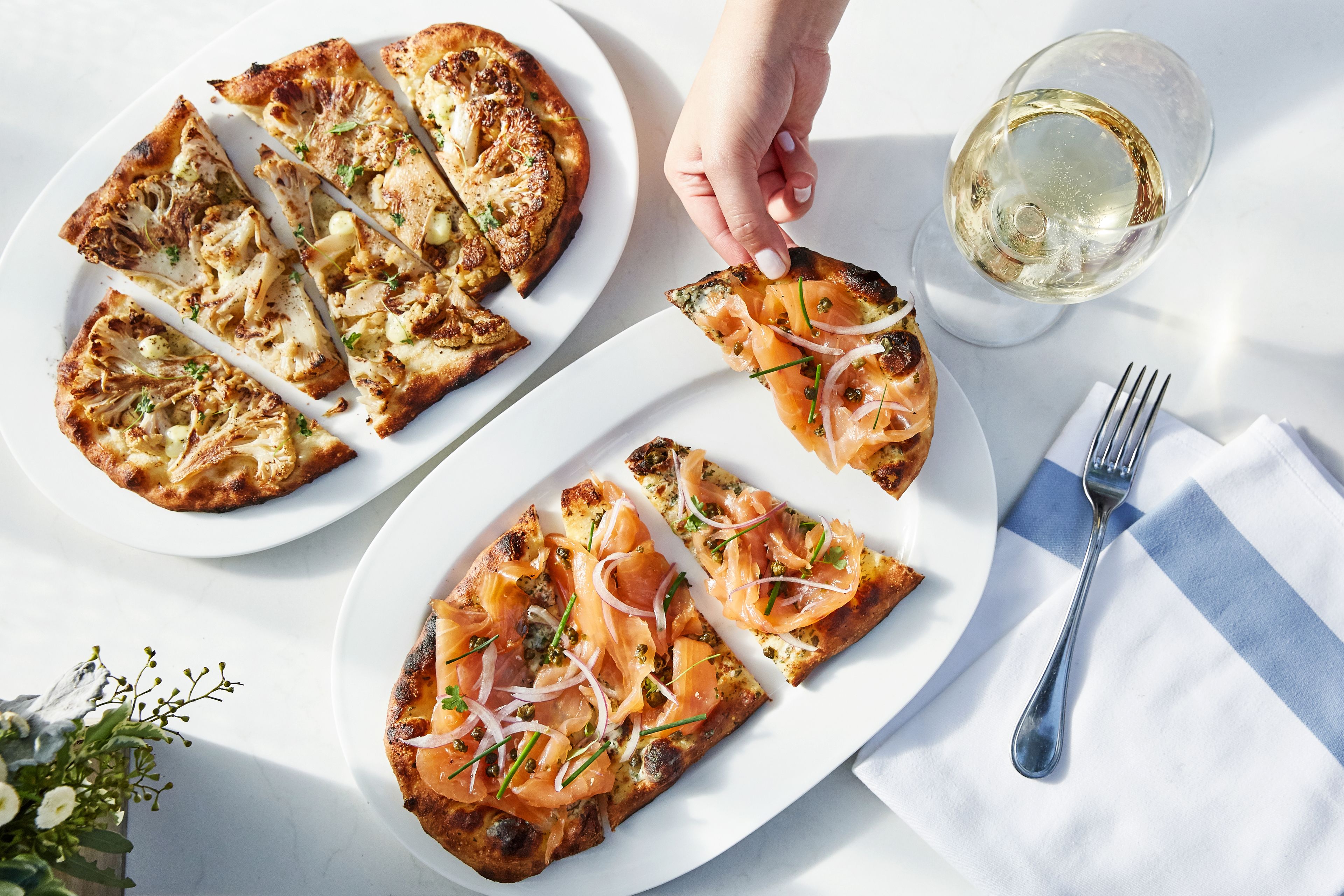 A table with two plates of pizza, a glass of white wine, and a fork on a white tablecloth.