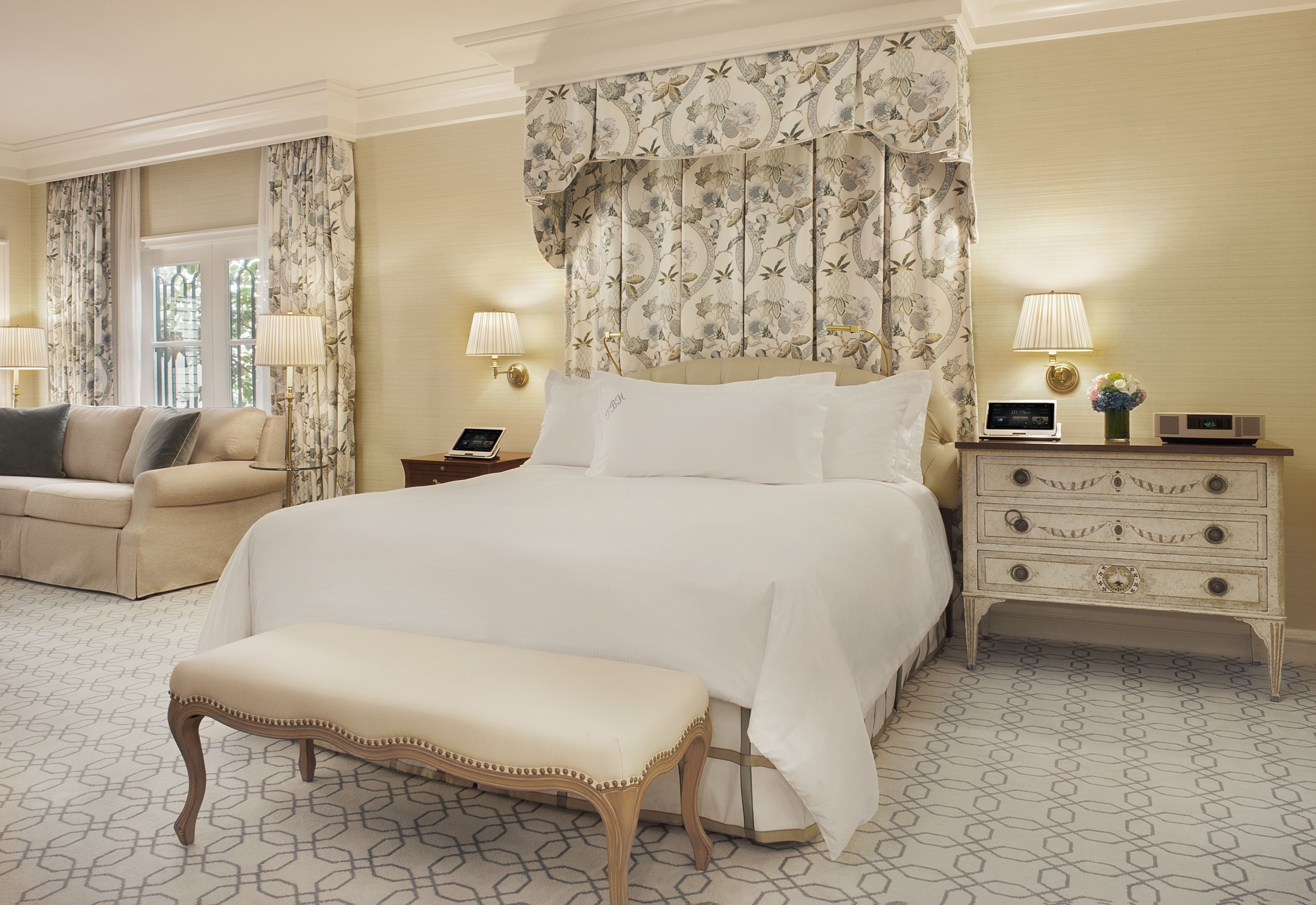 A hotel room with a large bed, a bedside table with a lamp, a nightstand, a bench, and a sofa.