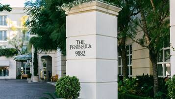 Un pilar blanco con la inscripción 'THE PENINSULA 9882', rodeado de follaje y un edificio al fondo.