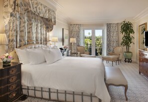 A hotel room with a large bed, two chairs, a desk, a dresser, and a large window with a view of greenery.