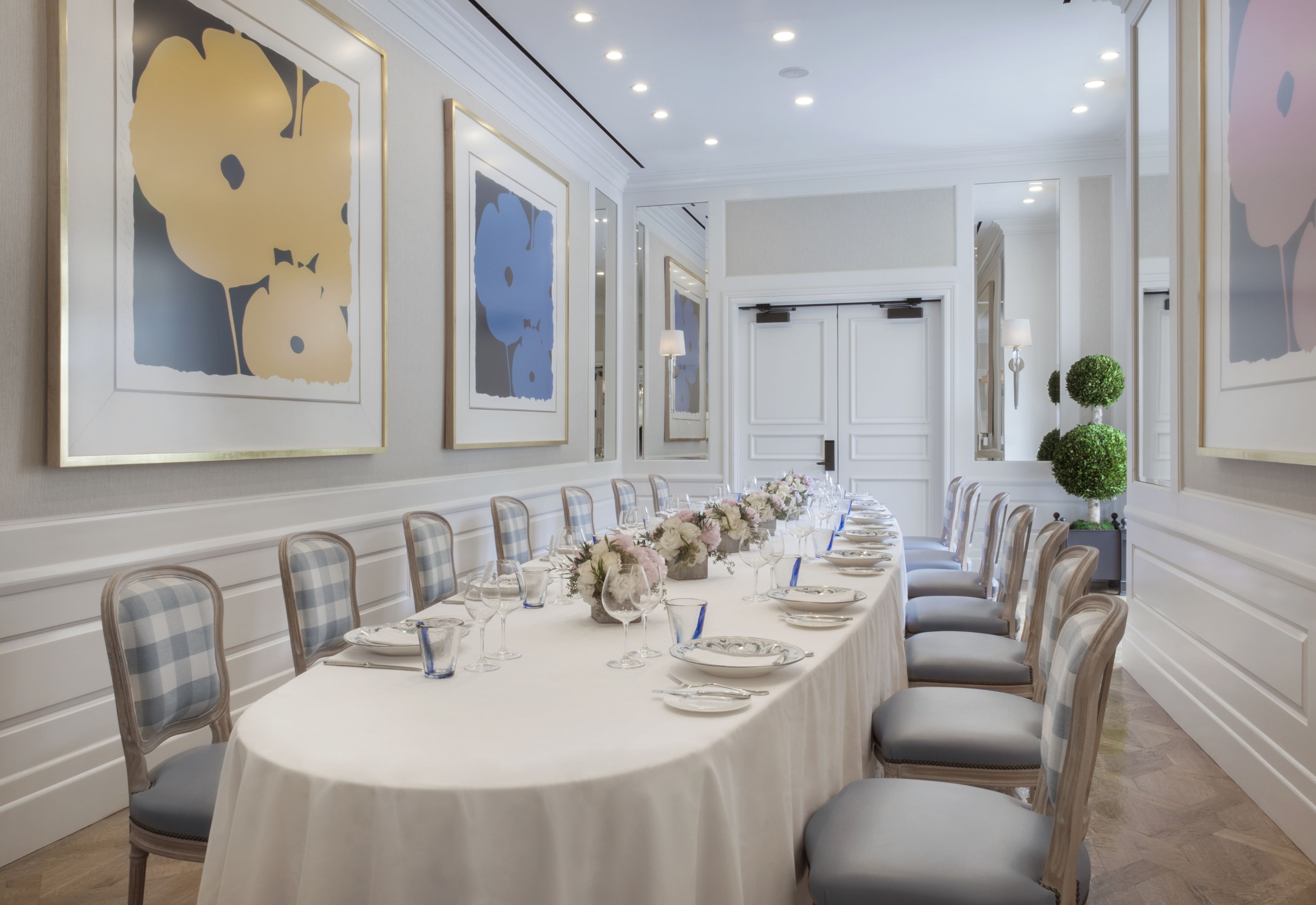 A long dining table set for a formal event, flanked by chairs with checkered upholstery, in a room with white walls and framed artwork.