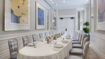 A long dining table set for a formal event, flanked by chairs with checkered upholstery, in a room with white walls and framed artwork.