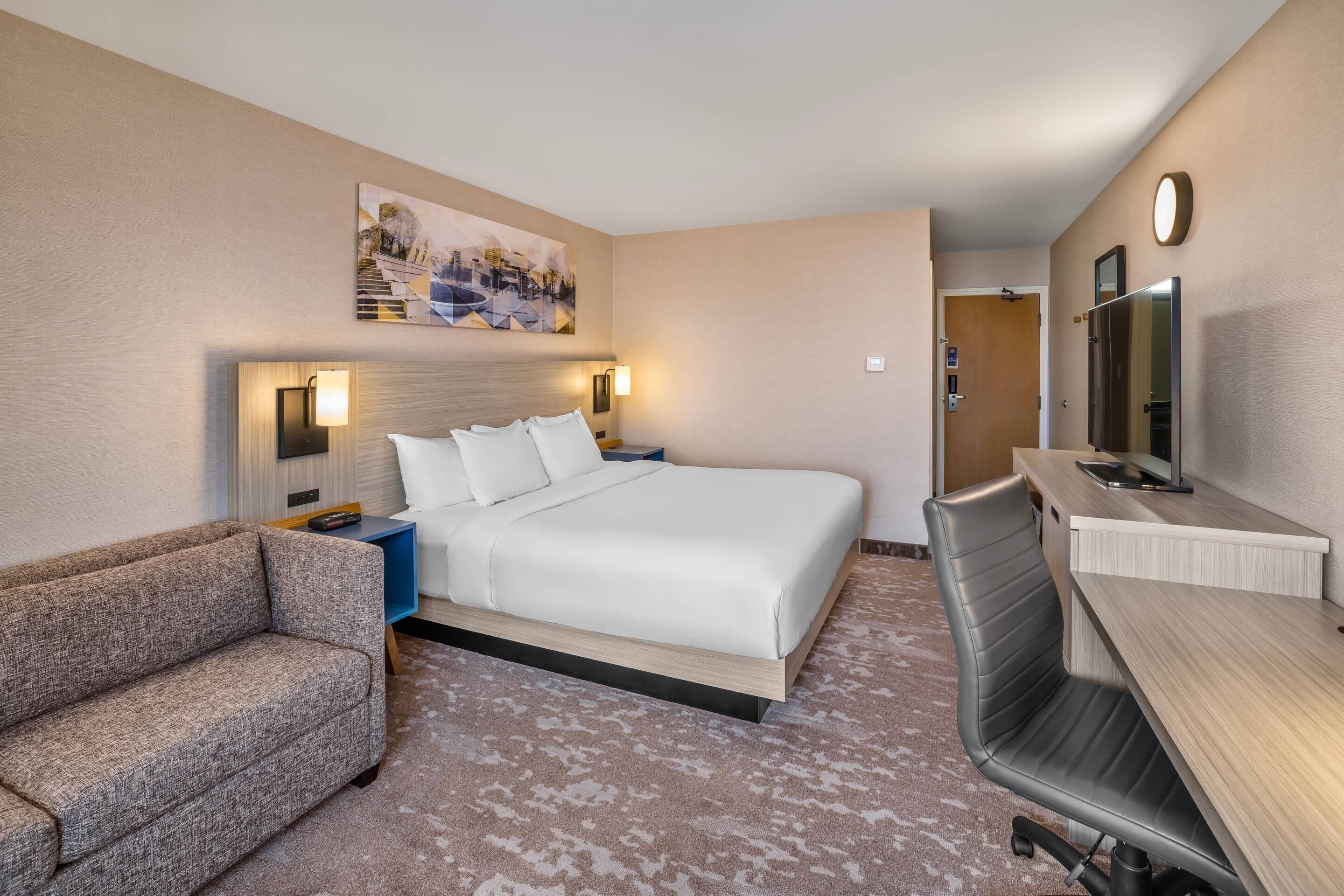 Une chambre d’hôtel avec un lit, un bureau, une chaise, un canapé et un tableau au mur.