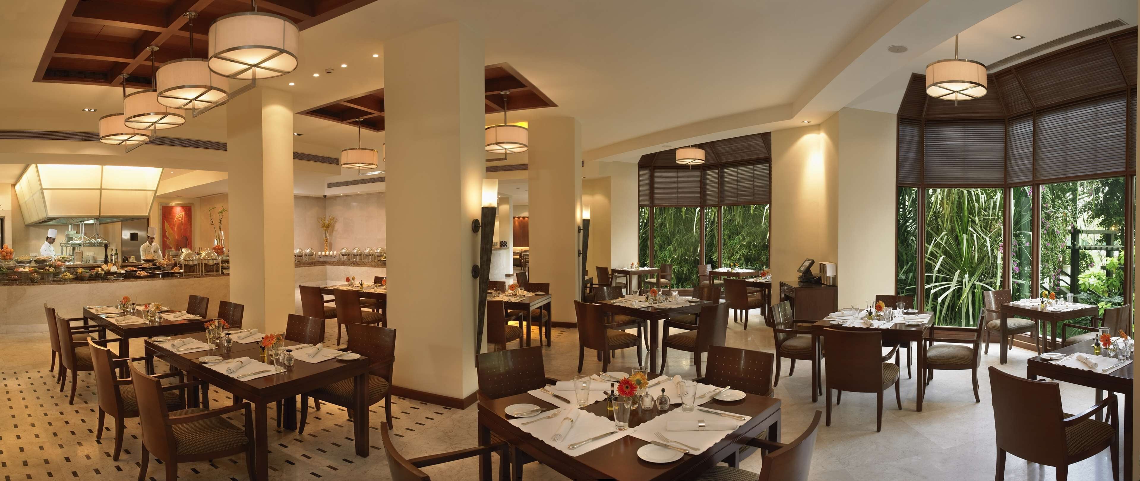 A spacious dining area with multiple tables and chairs, large windows with a view of greenery, and modern pendant lighting.
