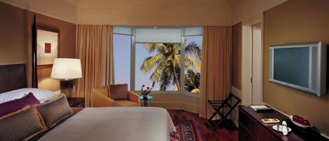 A hotel room with a bed, a chair, a TV, a desk with a laptop, and a window with a view of palm trees.