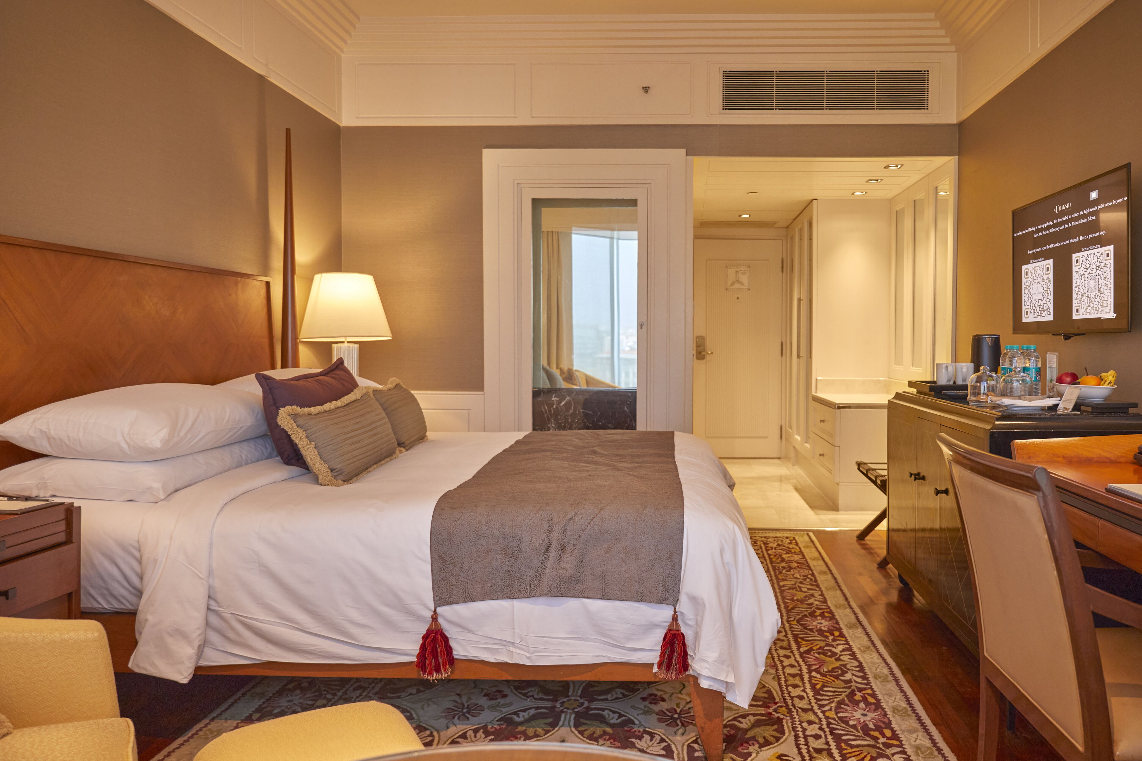 A hotel room with a large bed, a desk, a chair, a lamp, and a view of the city through the window.