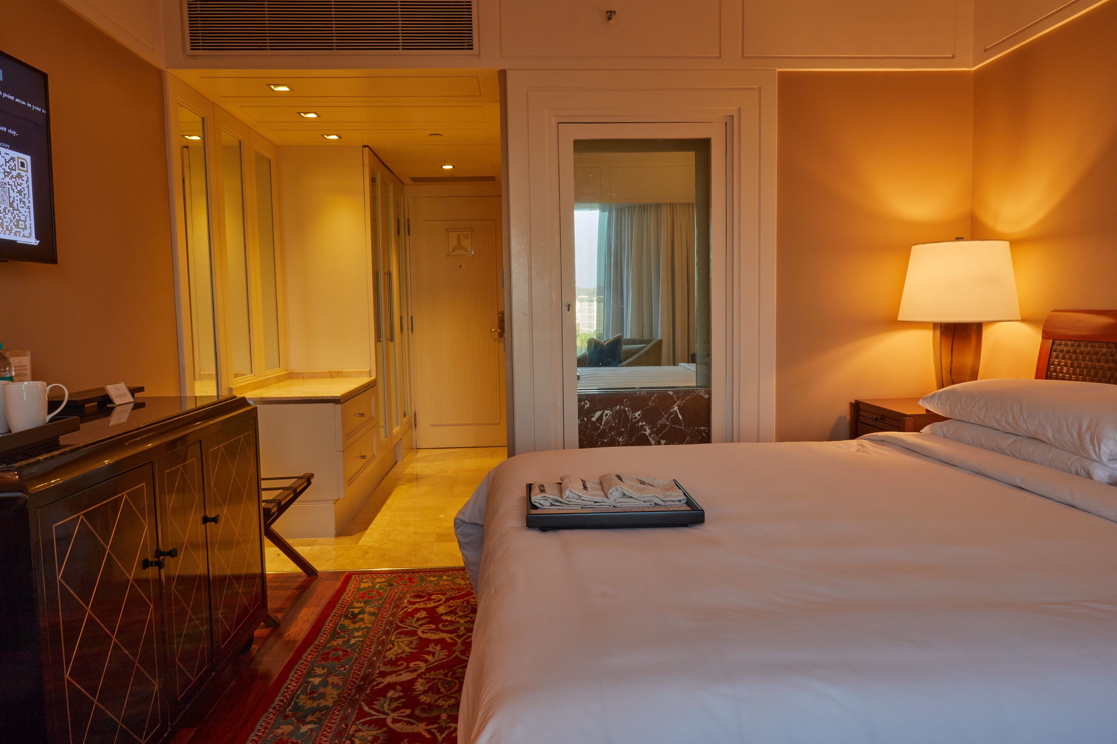 A hotel room with a neatly made bed, a bedside lamp, a dresser, and a view of the balcony through the door.