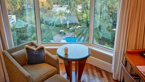 Habitación de hotel con un ventanal que ofrece vistas a una piscina y abundante vegetación. Hay una mesa redonda de cristal con una taza, y una silla acolchada con un cojín.