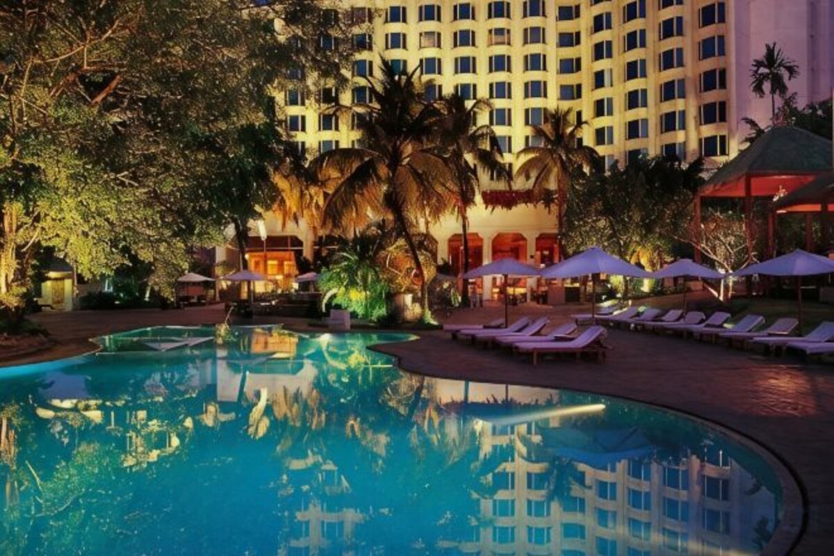 Una piscina de hotel con sillas de descanso y sombrillas, rodeada de palmeras y un edificio de varios pisos.