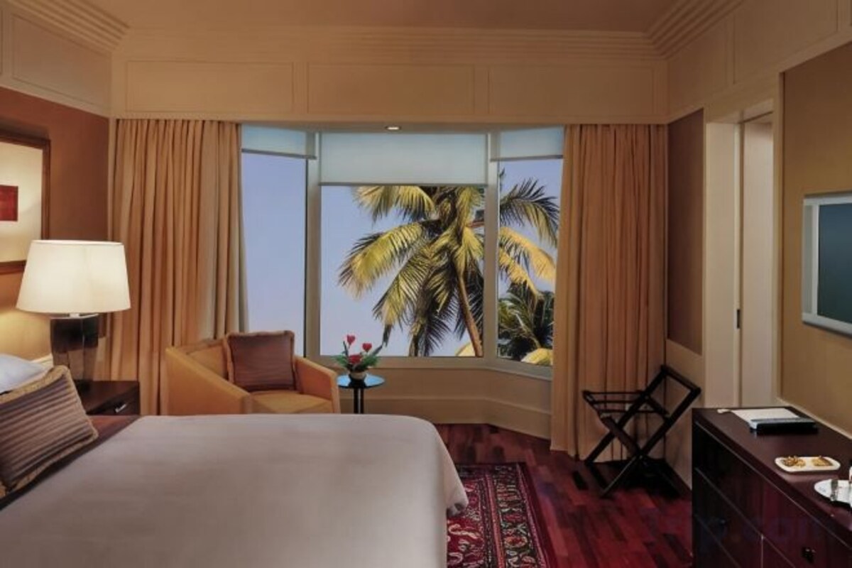 Habitación de hotel con una cama grande, un escritorio, una silla, una mesita con flores y vista a palmeras a través de una ventana.