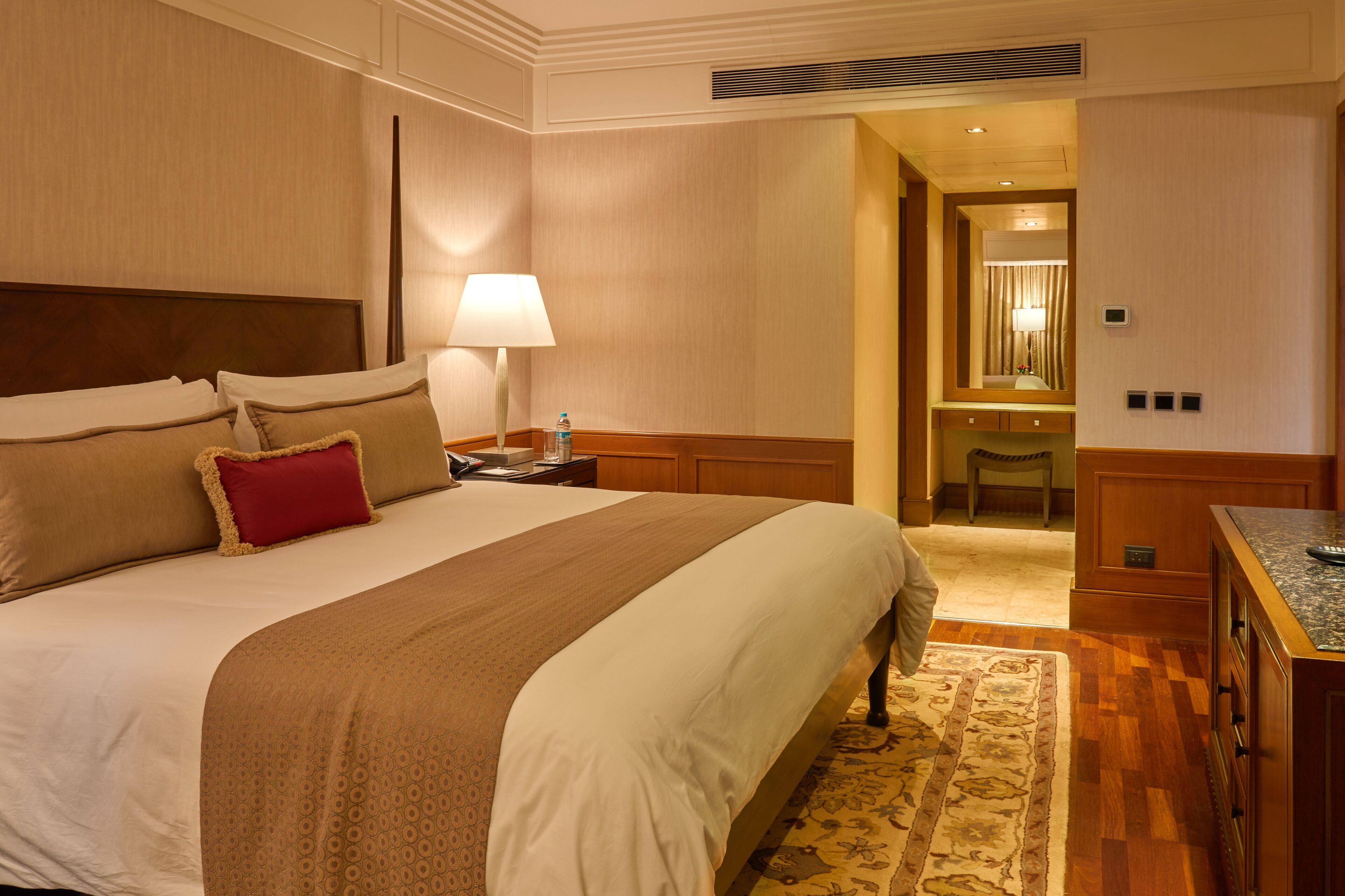 Una habitación de hotel con una cama grande, una mesita de noche con lámpara, cabecera de madera, baño con espejo y un escritorio pequeño con cajones.