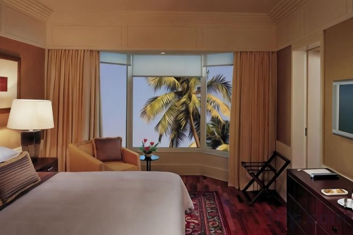 Habitación de hotel con una cama grande, un escritorio, una silla, una mesita con flores y vista a palmeras a través de una ventana.