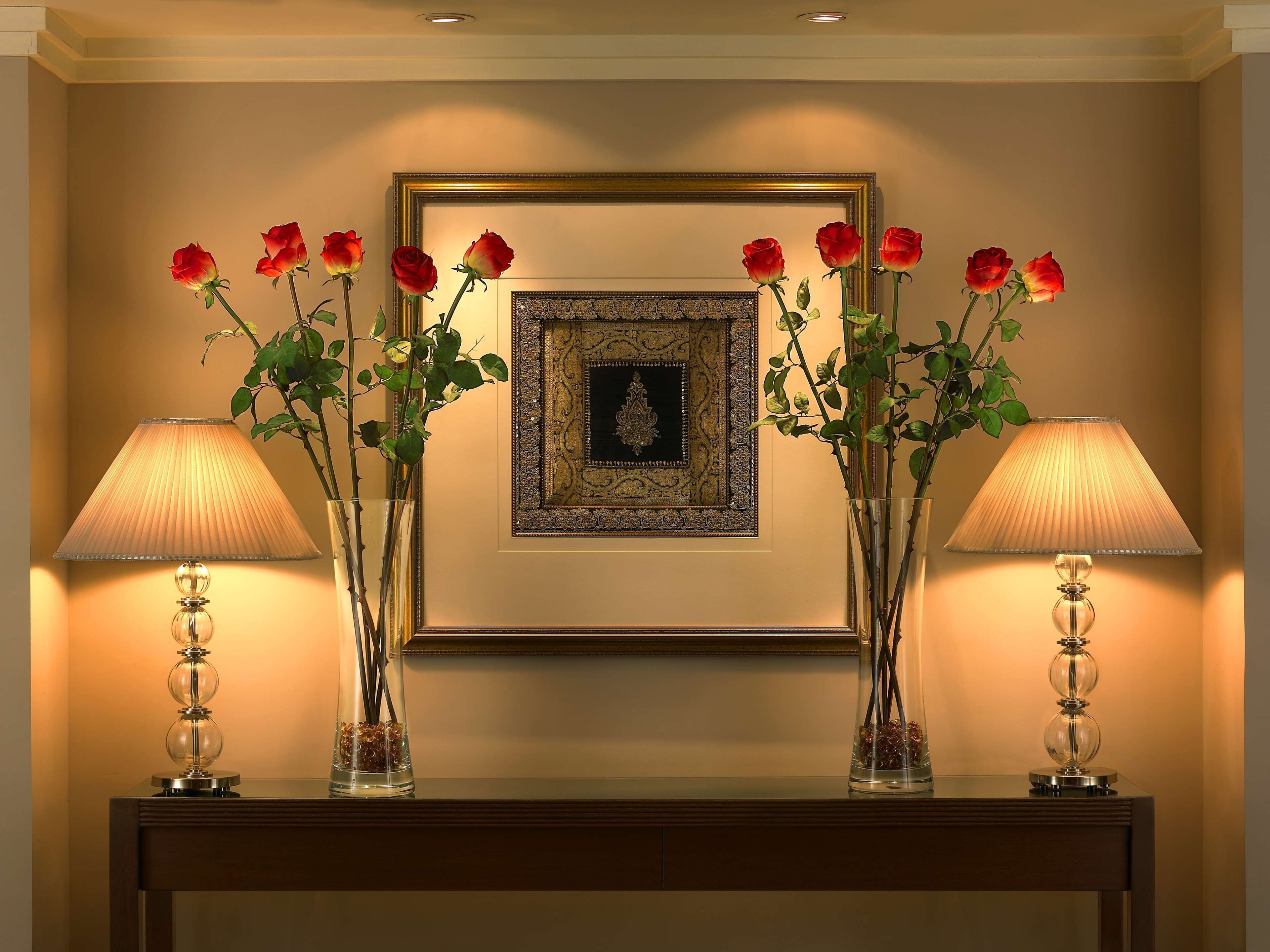 A framed artwork above a console table with two lamps and vases of red roses.
