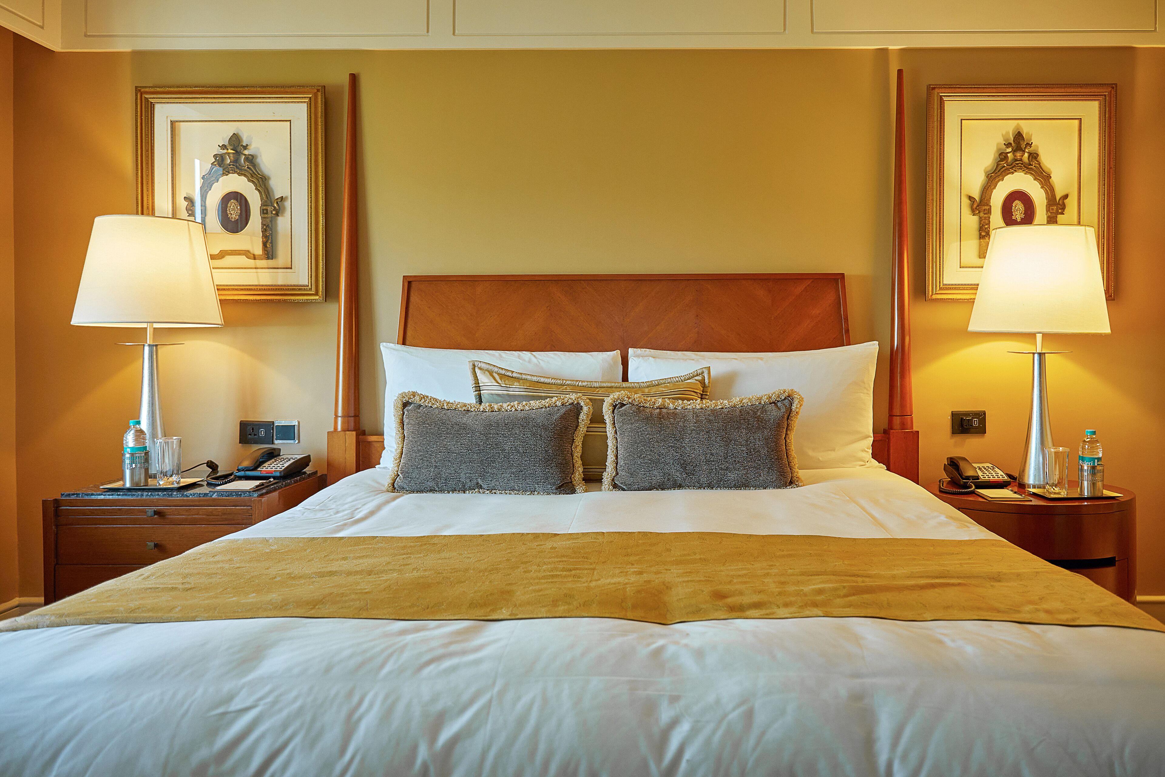 Habitación de hotel con una cama grande, dos mesitas de noche, dos lámparas, un teléfono, una botella de agua y cuadros en las paredes.