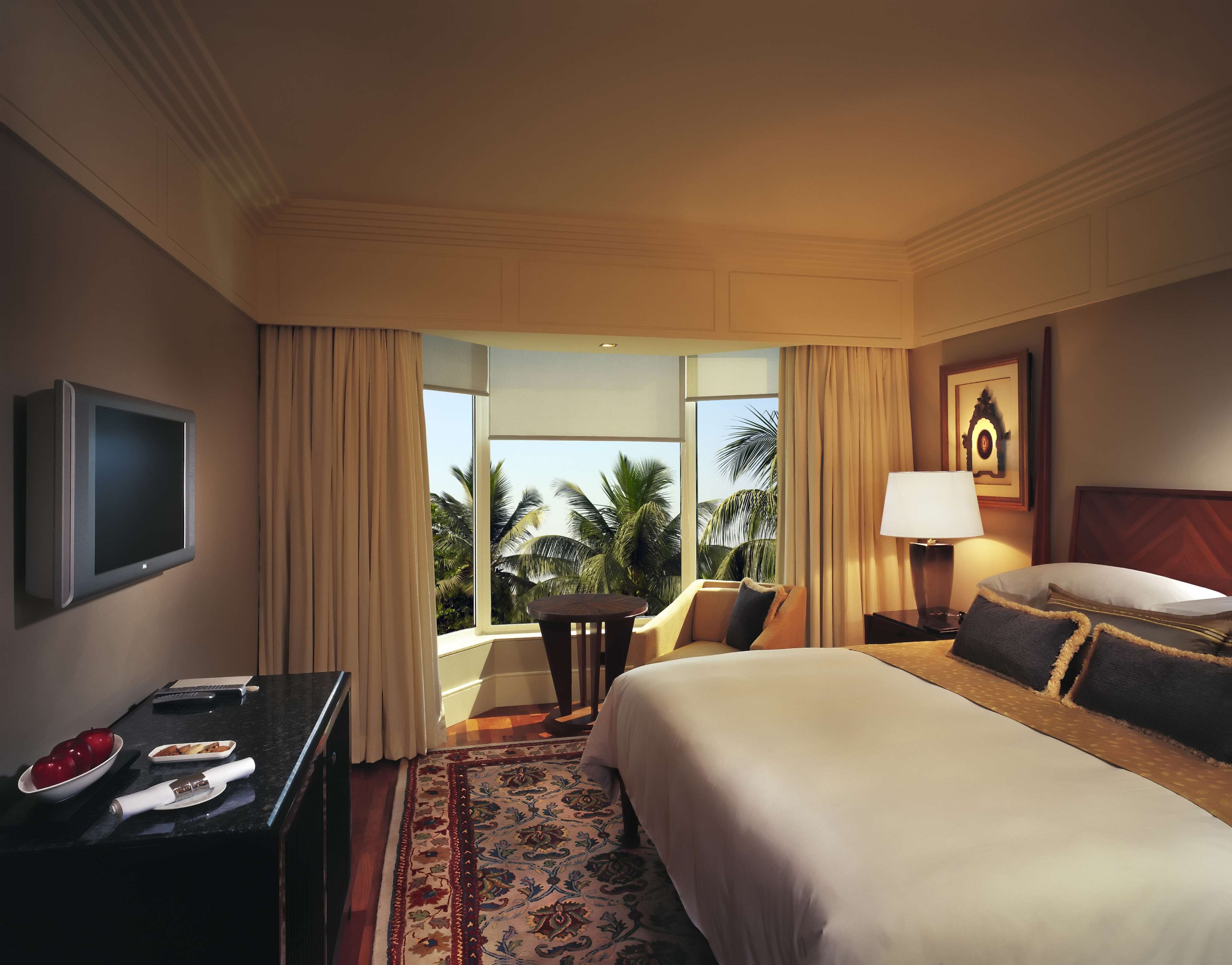 A hotel room with a large bed, a desk with a TV, a lamp, and a view of palm trees.
