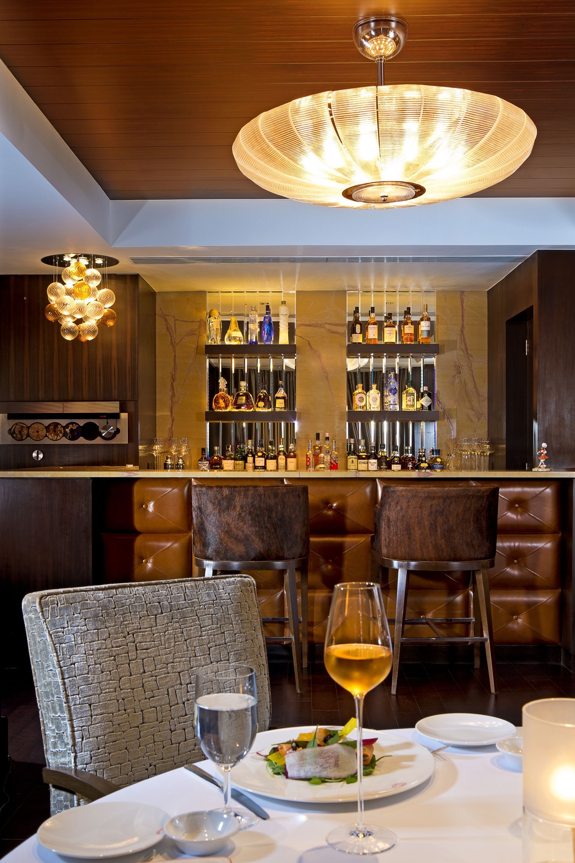 Un'area bar con soffitto in legno, un grande lampadario e una profonda selezione di liquori. Sono presenti sgabelli da bar in pelle e un tavolo da pranzo con un piatto di cibo e un bicchiere di vino.