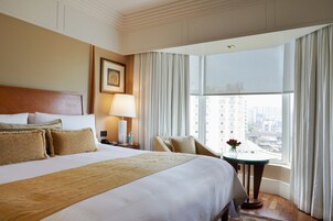 A hotel room with a large bed, a sitting area with a chair, a small table with a vase of flowers, and a window with curtains.