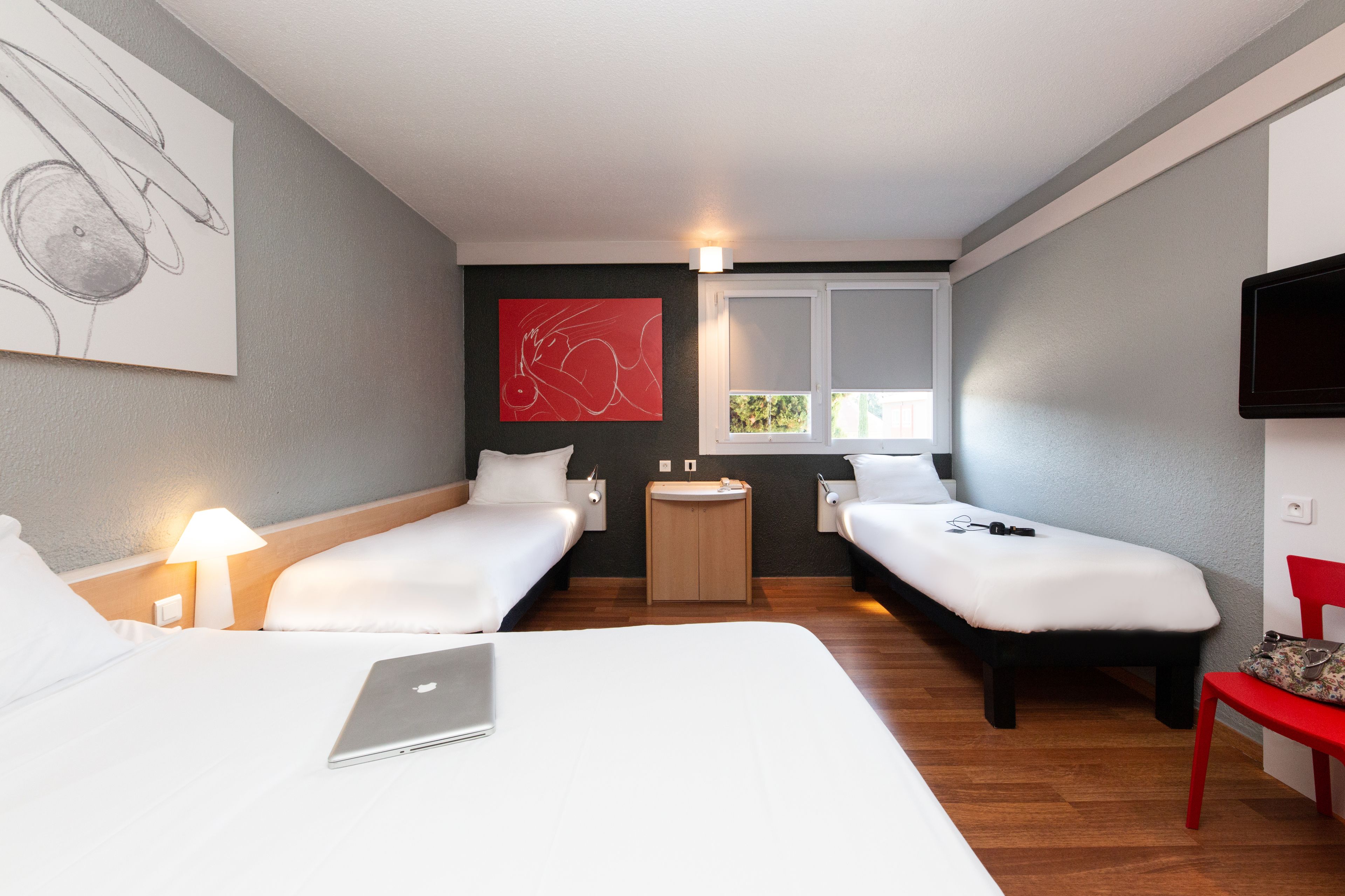 A hotel room with two single beds, a desk with a laptop, a small round table with a lamp, a red chair, and a TV mounted on the wall.