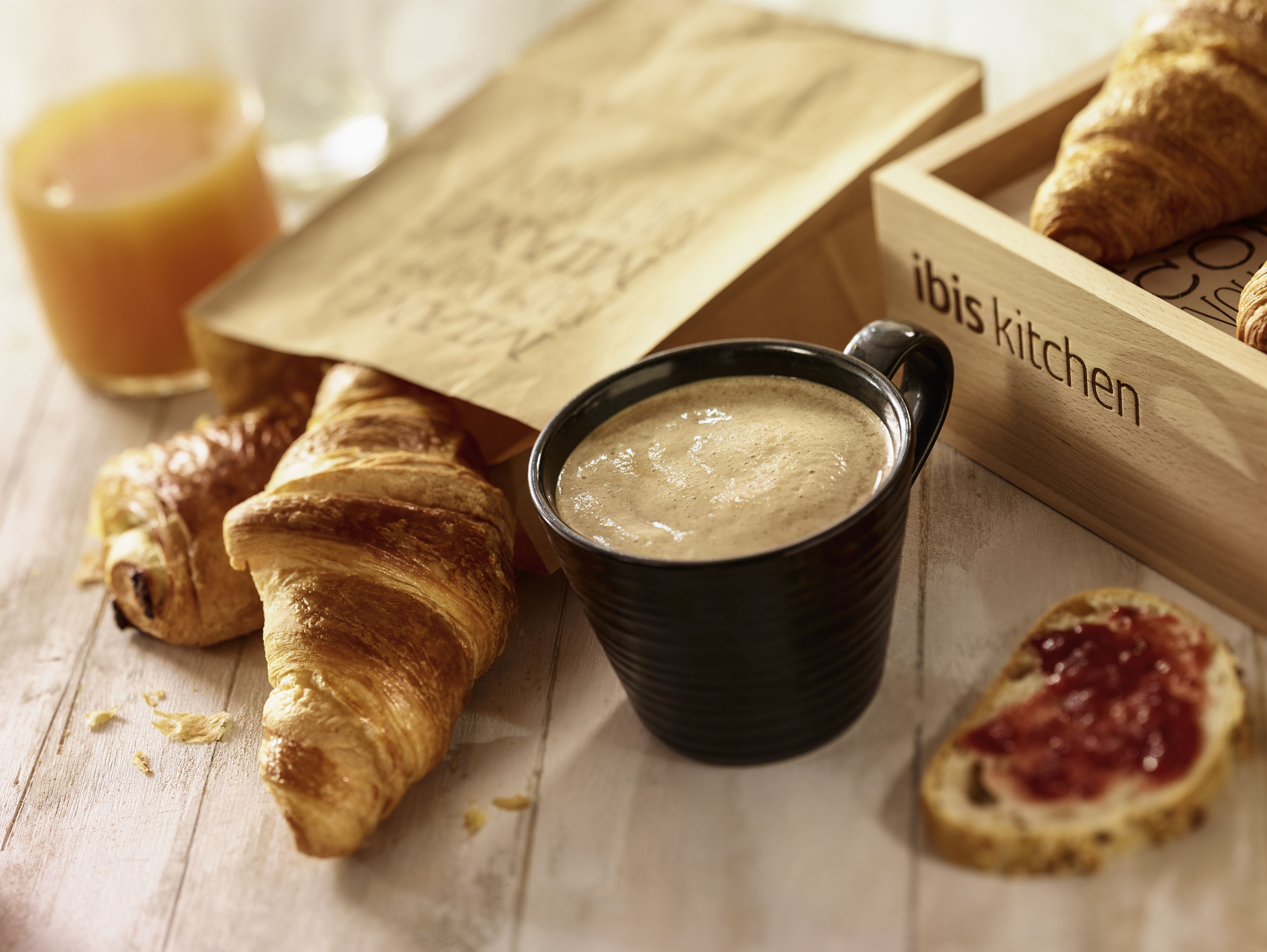 A wooden tray with croissants, a black mug with a creamy beverage, and a paper bag with text.