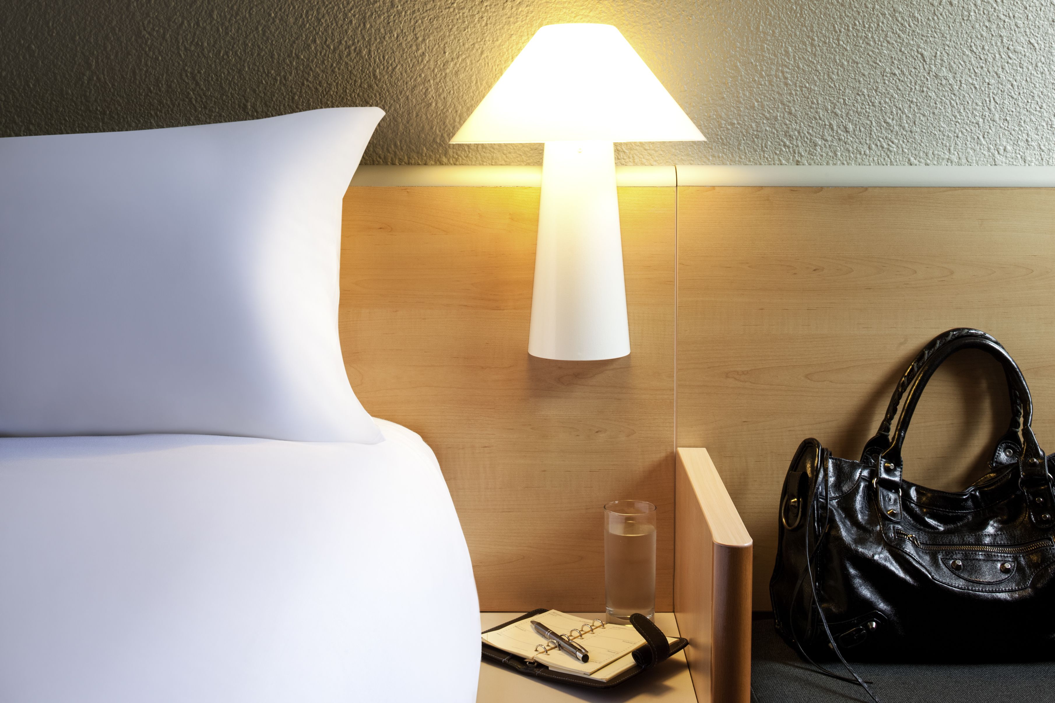 Una cama bien hecha con una almohada blanca, una lámpara de noche, un vaso de agua, un cuaderno y un bolso negro sobre una mesita de noche de madera.