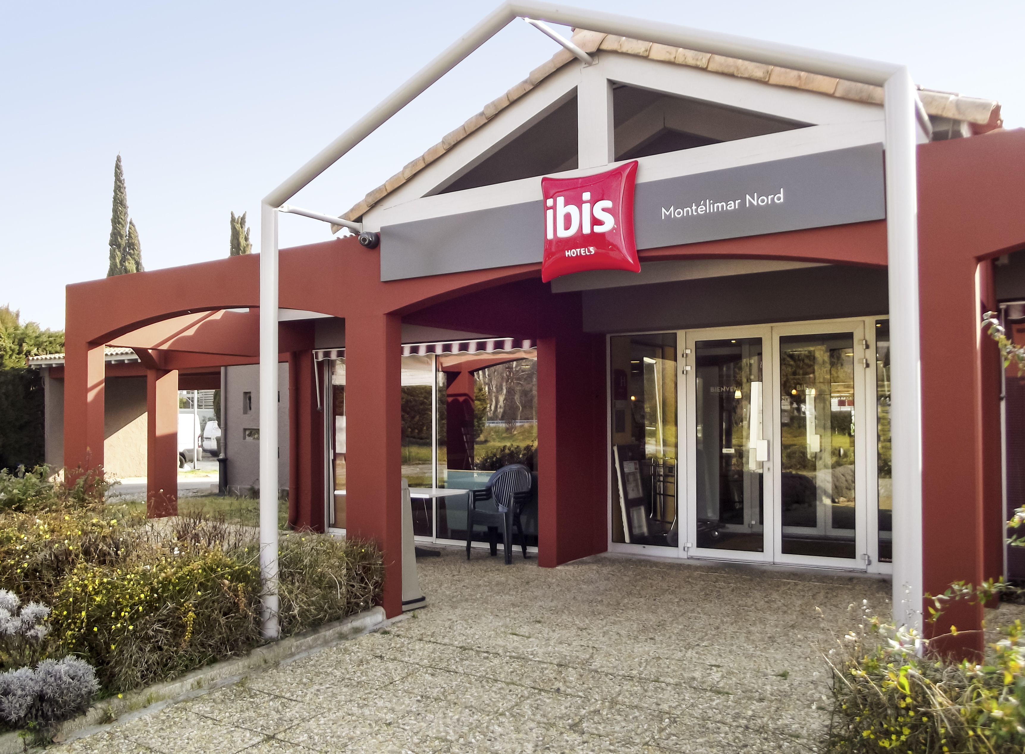 A modern hotel entrance with a red "ibis" sign, glass doors, and a covered outdoor seating area.