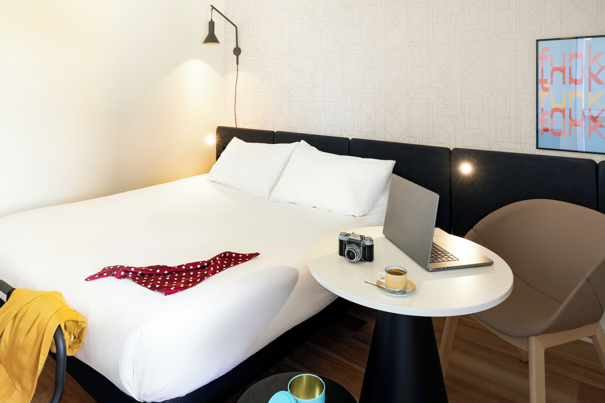 A hotel room with a bed, a round table with a laptop, a camera, and a cup.